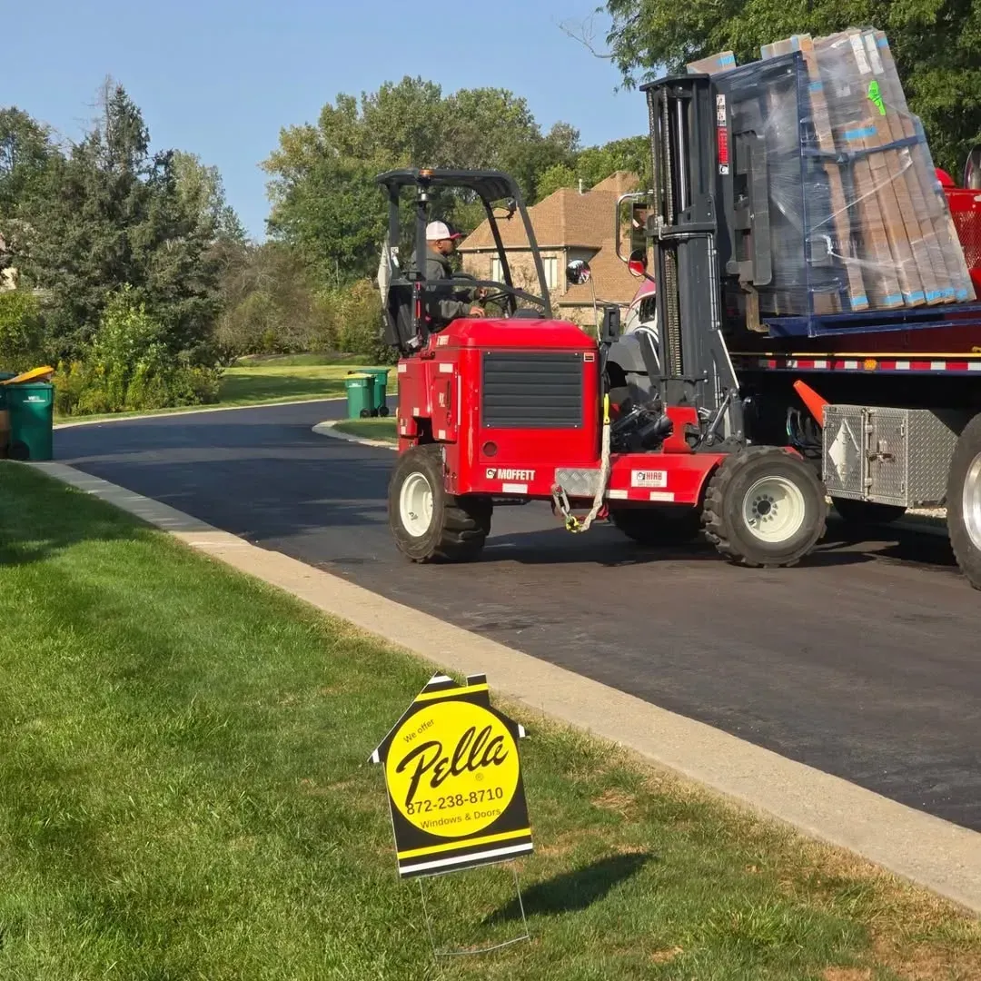 Red pavement roller beside a truck on a suburban road, with a yellow yard sign in the foreground.