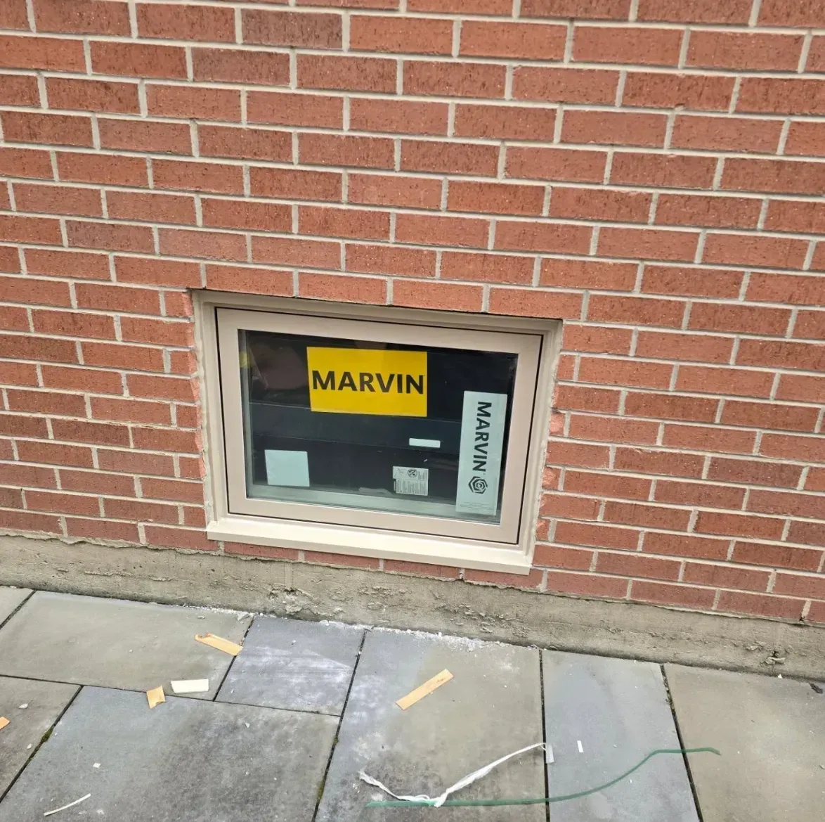 Small basement window in a brick wall with Marvin labels and a sidewalk below.
