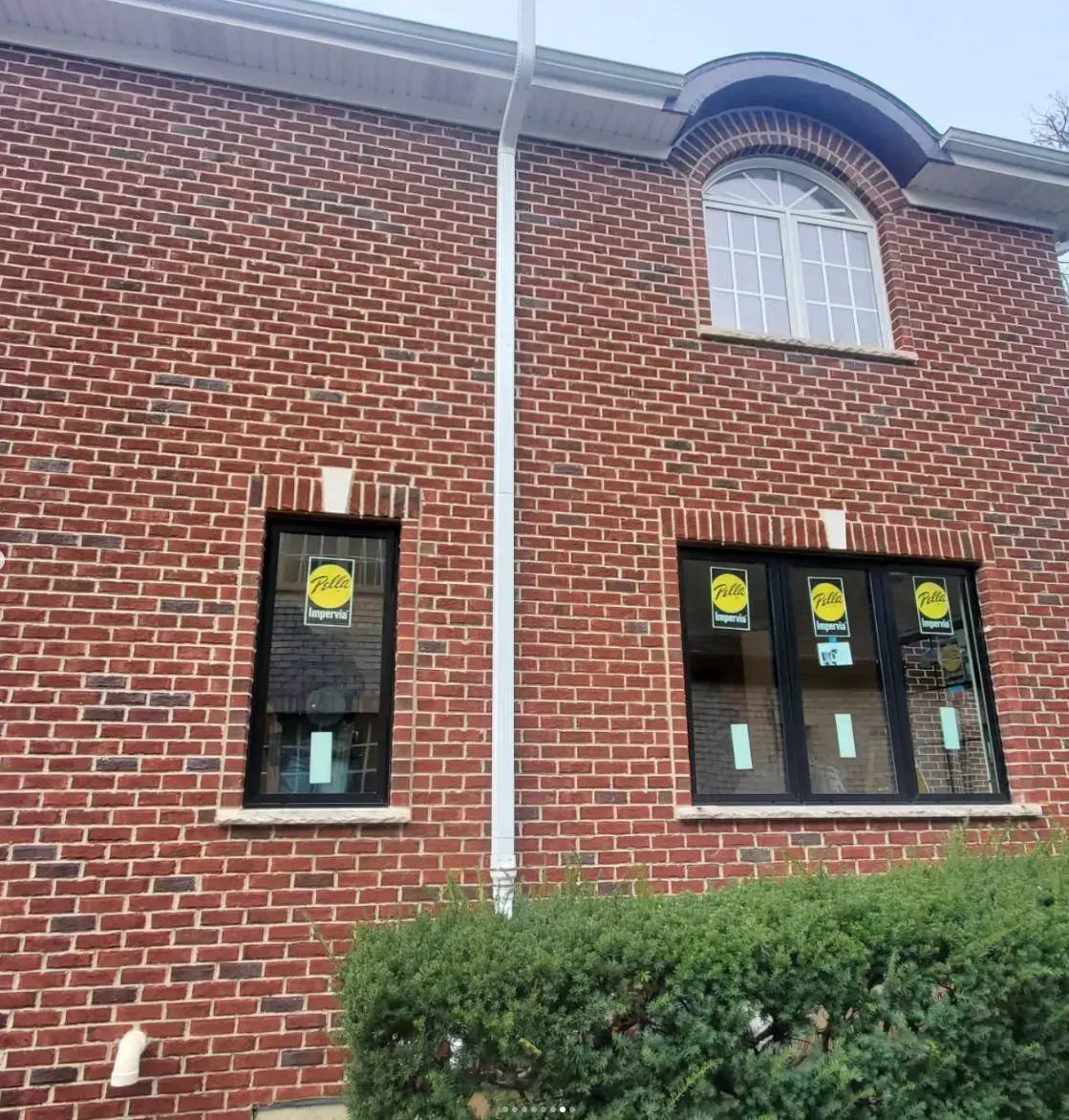 Brick building facade with small windows, white trim, and green shrubs in front.