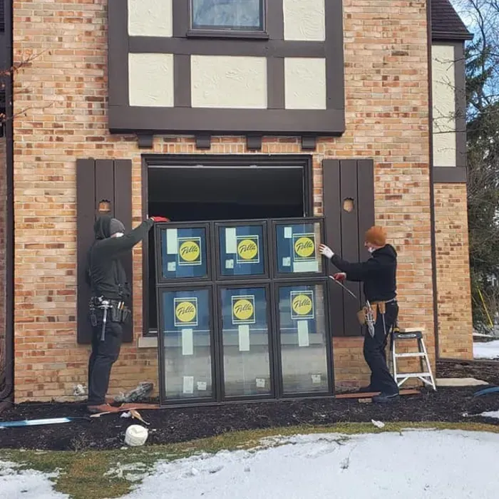 Two workers install large window panels on a brick house under construction.