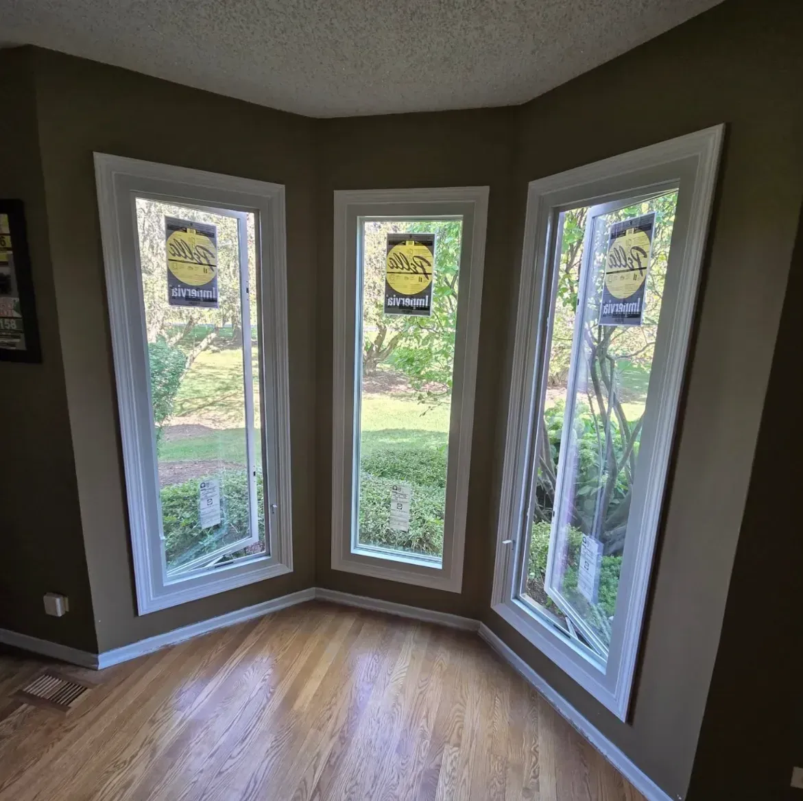 Bay window nook with three tall windows, hardwood floor, and a green backyard view.