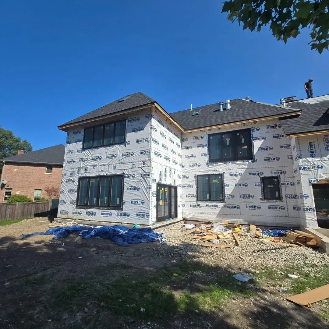 Two-story house under renovation, wrapped in white housewrap with exposed siding and debris in front.