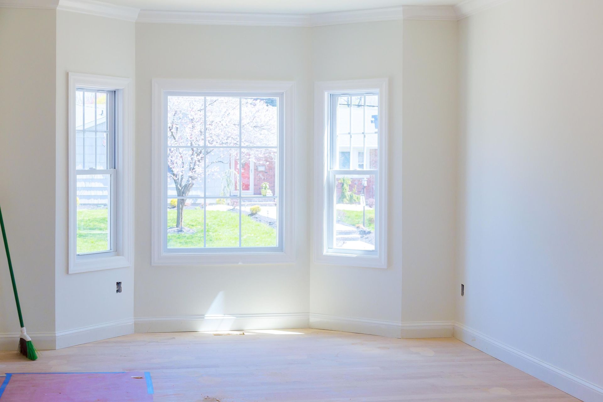 Bright empty room with white walls, hardwood floor, and three tall windows overlooking a green yard