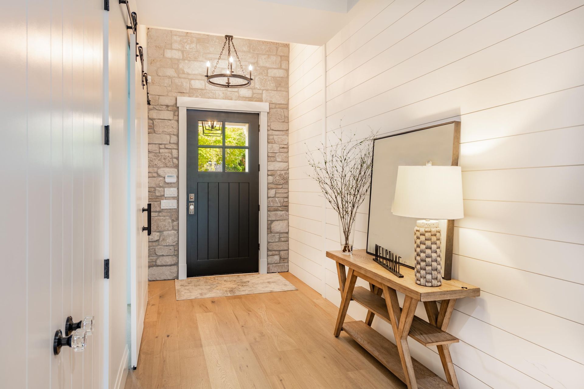 Bright entry hallway with black front door, light wood floors, and rustic bench with lamp.