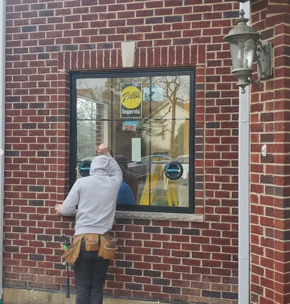 Person in gray hoodie standing at a brick storefront window with a hanging lantern.