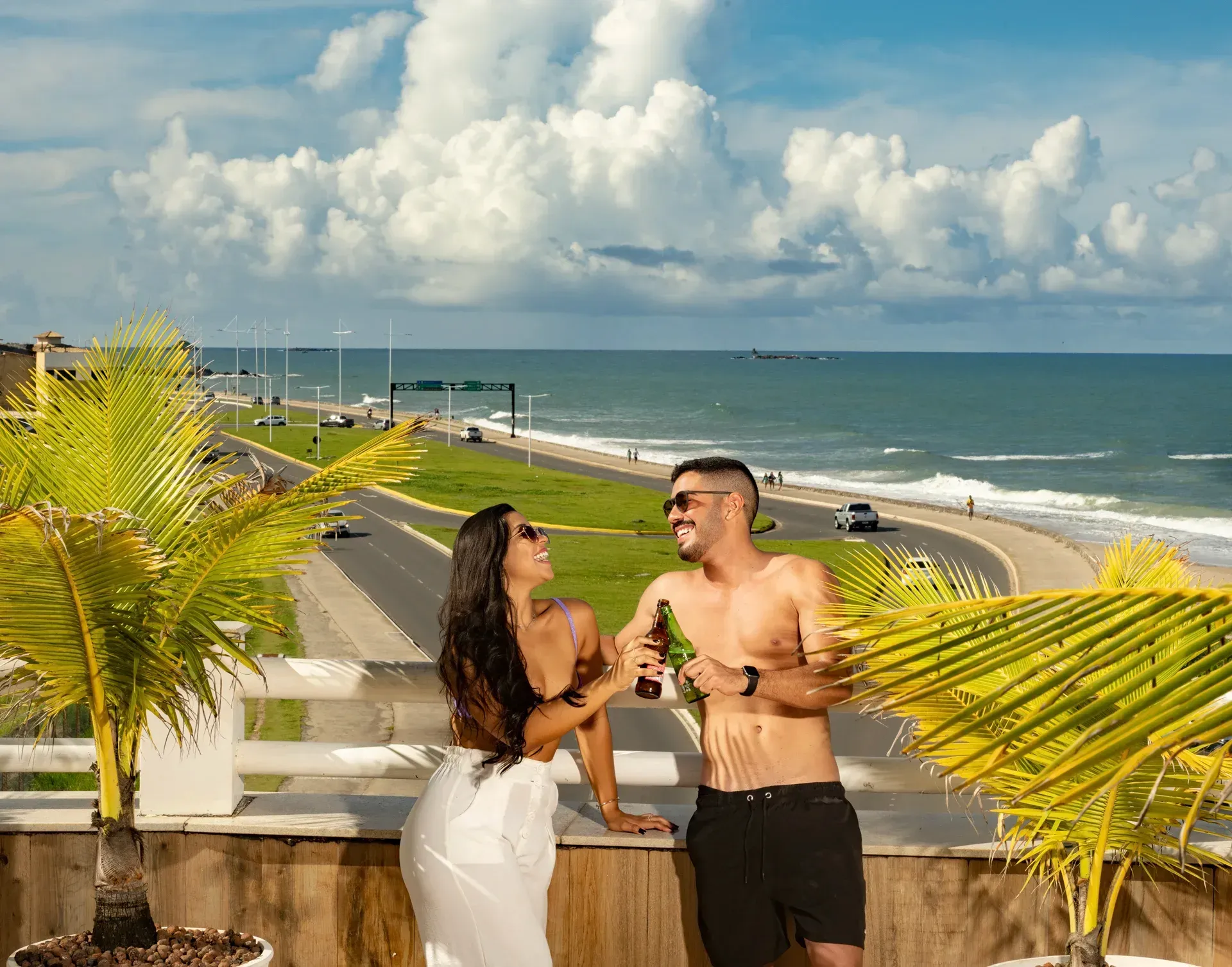 Um homem e uma mulher bebem cerveja em uma varanda com vista para o mar.