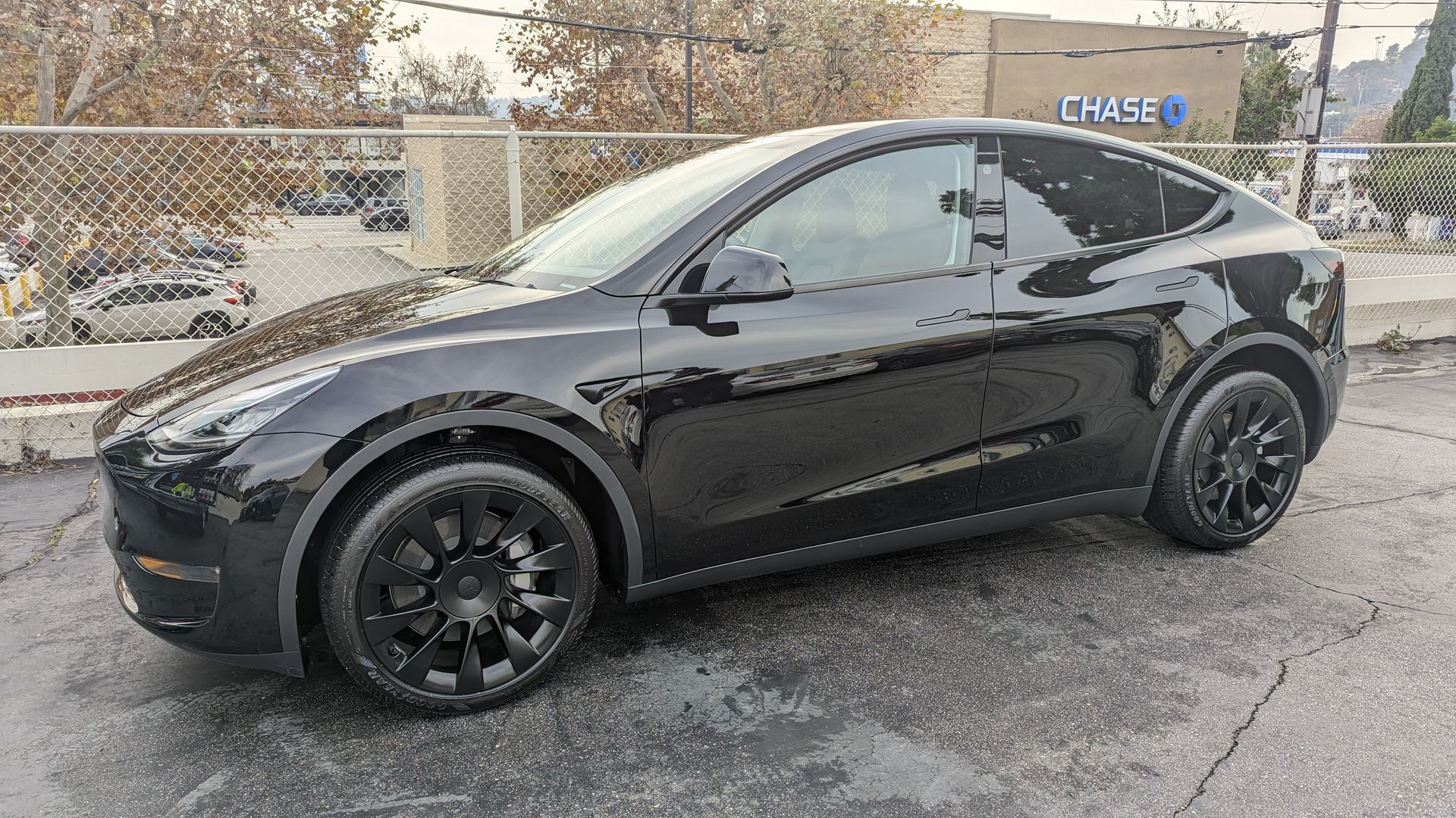 Black Tesla parked on a wet lot beside a chain-link fence with snow in the background