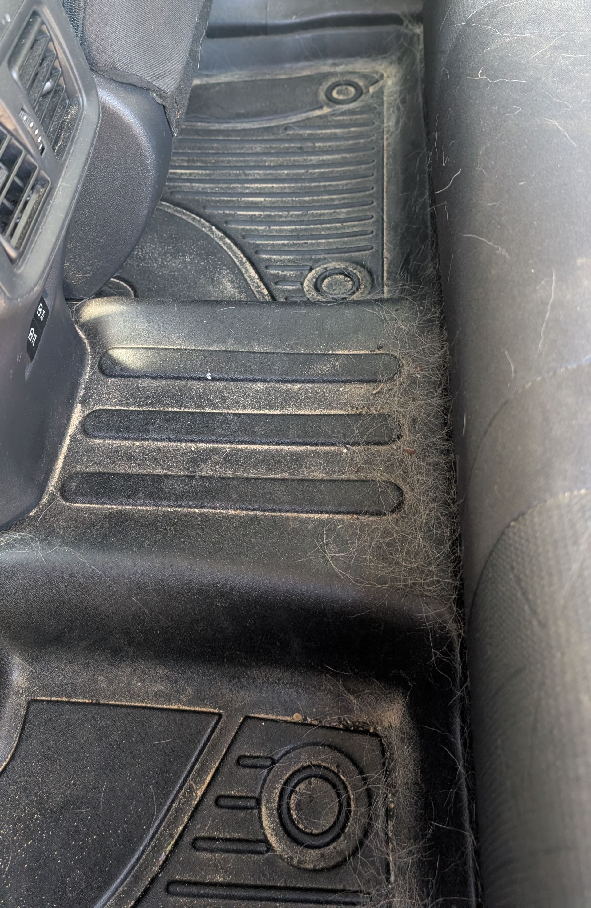 Car interior floor and center tunnel with black rubber mats and a vent panel near the seat.