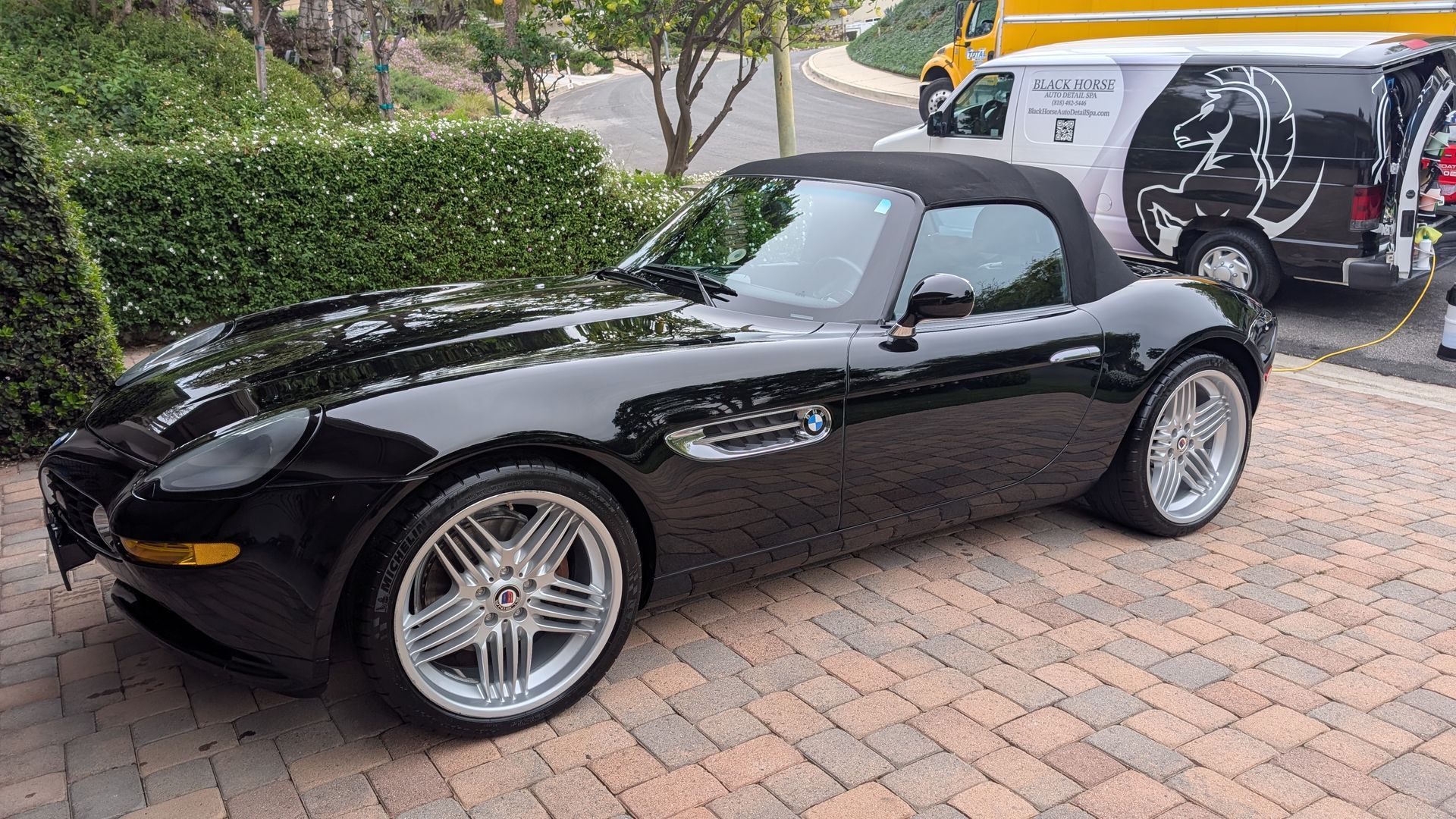 Black convertible sports car parked on a brick driveway beside a white van and hedge