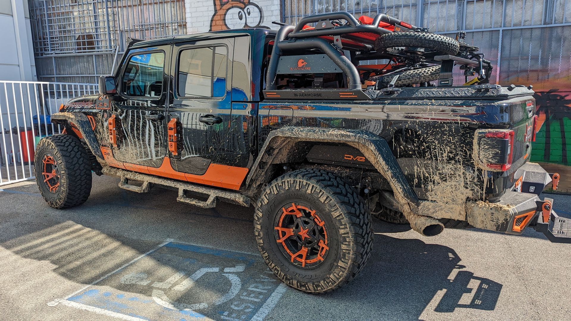 Mud-covered black and orange pickup truck with large off-road tires parked outdoors at a shop.