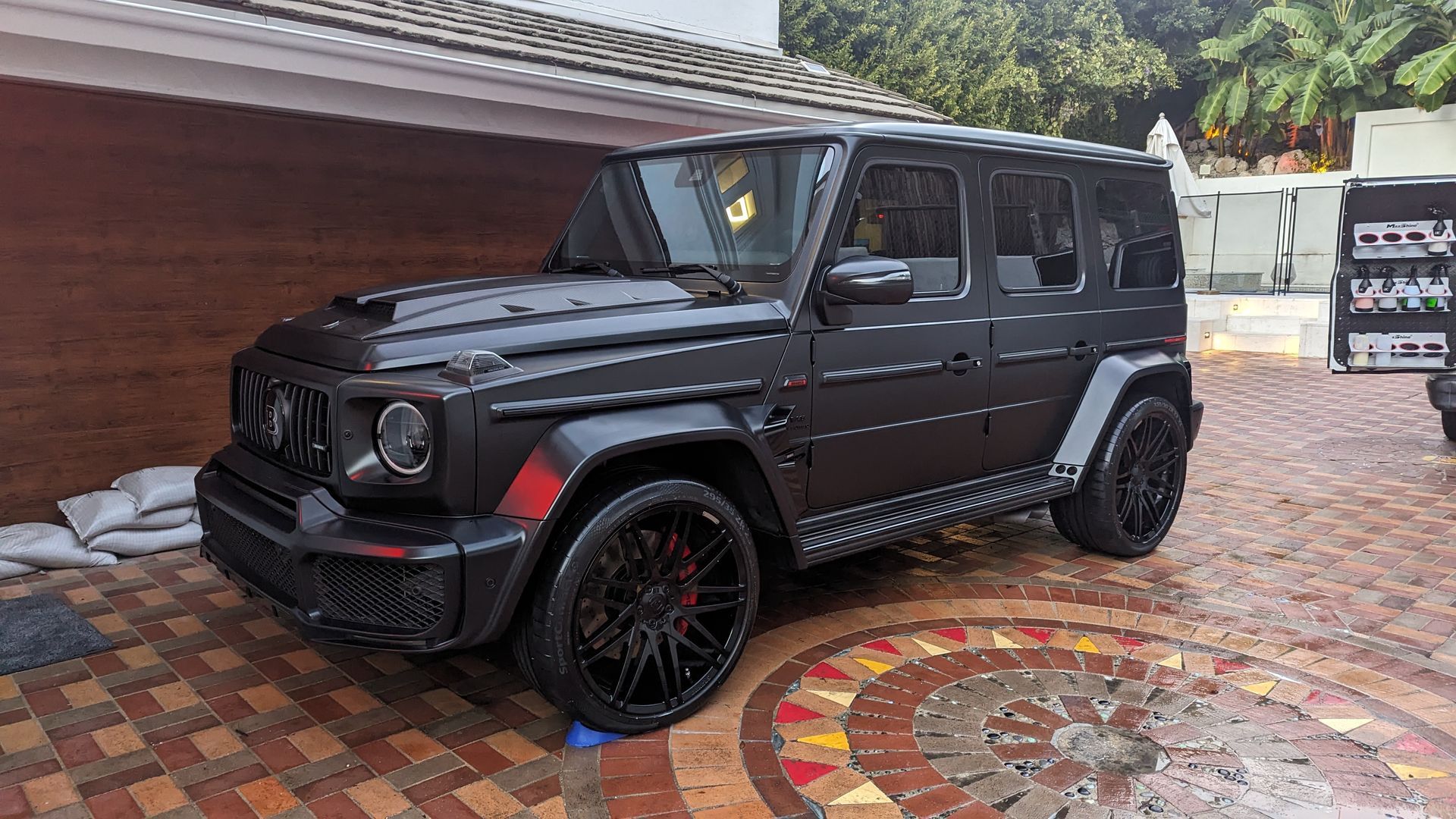 Black Mercedes G-Class SUV parked on a brick driveway beside a red building.