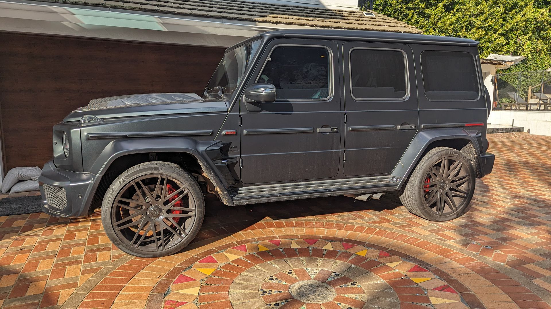 Dark gray Mercedes G-Class SUV parked on a patterned driveway in front of a house.