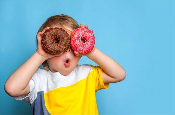 A person holding a chocolate donut over one eye and a pink frosted donut over the other against a blue background.