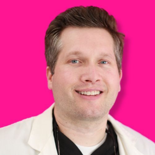 A smiling person with short, light brown hair wearing a white lab coat against a bright pink background.