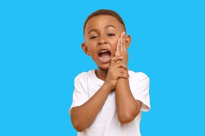 A child with an open mouth holds their hand against their cheek, expressing pain or discomfort against a blue background.