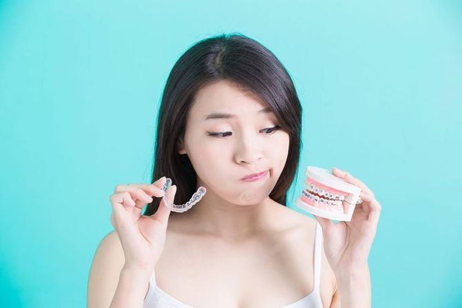 A person in front of a teal background holds a clear aligner tray in one hand and a model of teeth with braces in the other.
