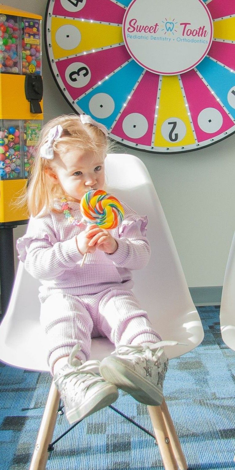 A toddler in a light purple outfit sits in a chair eating a colorful swirl lollipop in front of a game wheel.