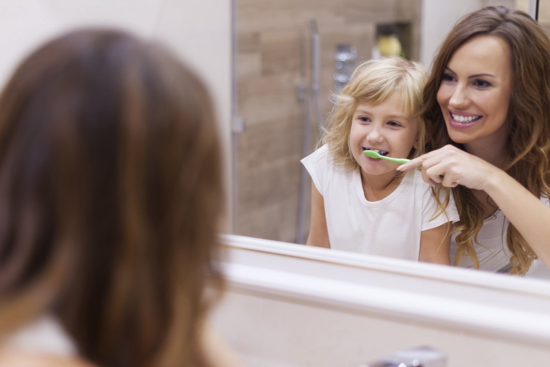 A smiling adult helps a young child brush their teeth in front of a bathroom mirror.
