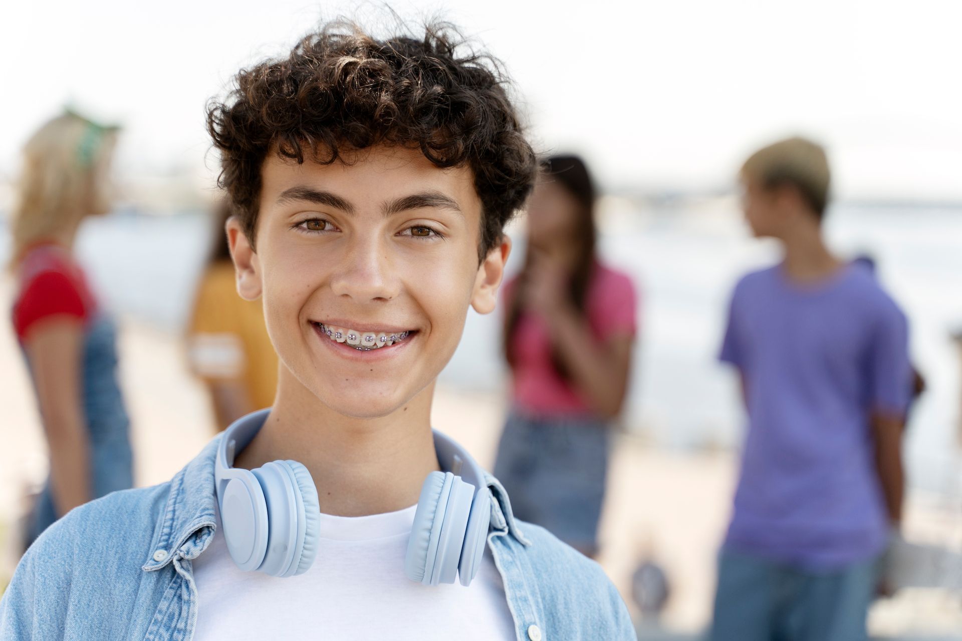 A smiling person with curly hair and braces wears a blue shirt and headphones around their neck, with others in the back.