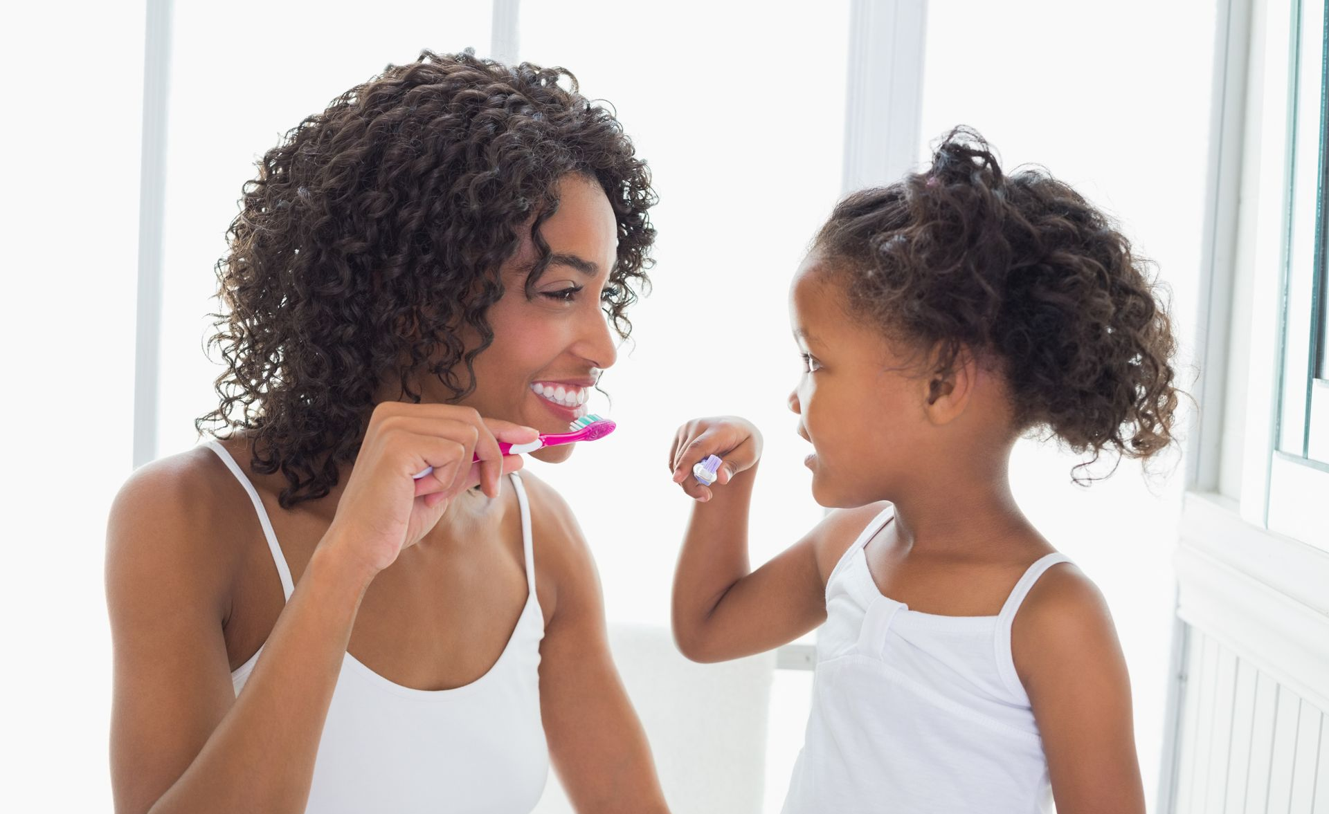 Two children in a bright bathroom, one brushing teeth while the other sits on the counter looking into a mirror.