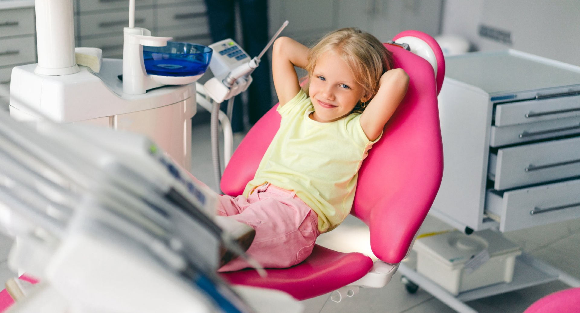A child with hands behind their head smiles while sitting in a pink dental chair inside an exam room.