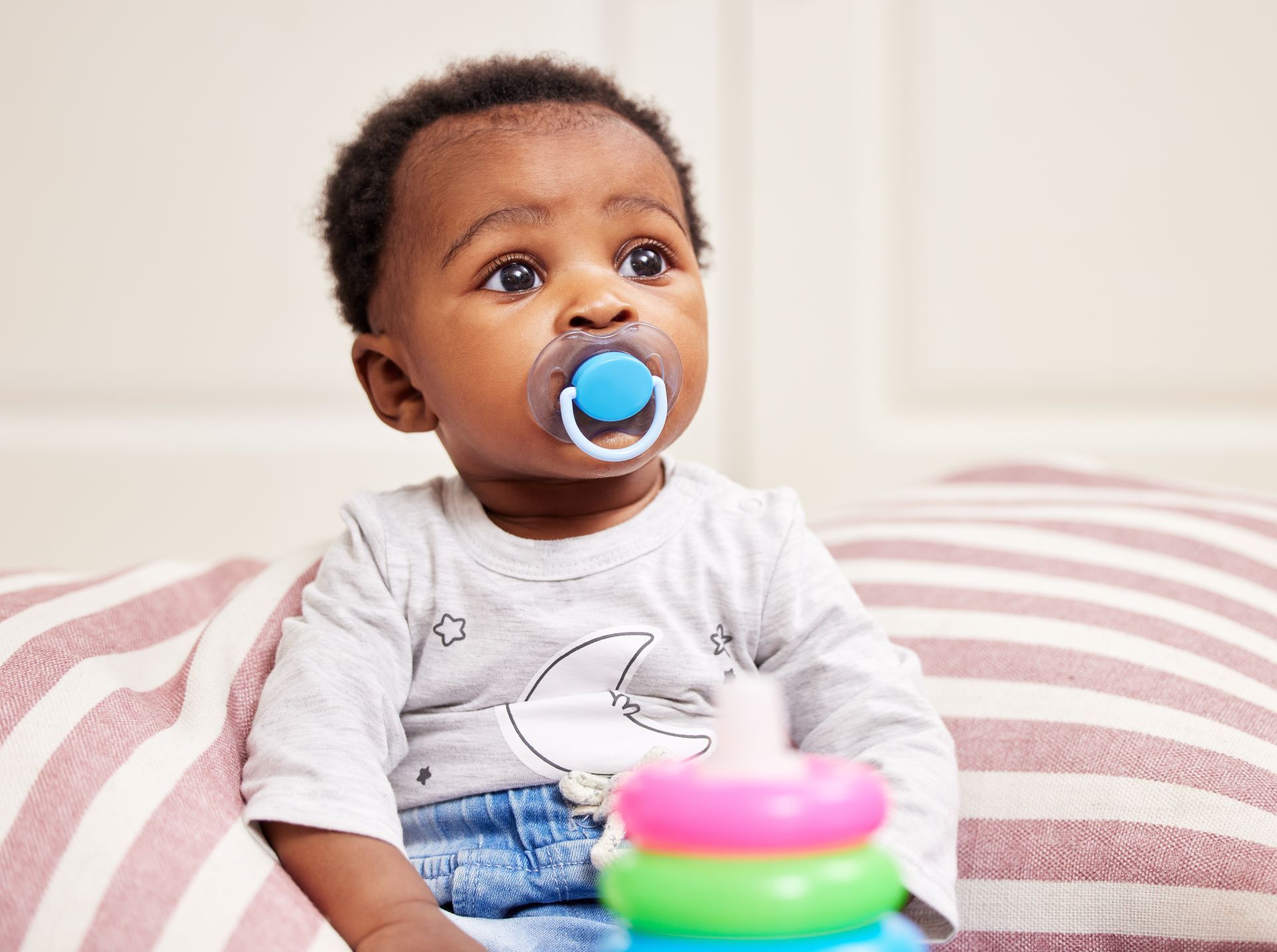 Baby with blue pacifier sitting on a striped cushion, reaching toward a colorful stacking toy.