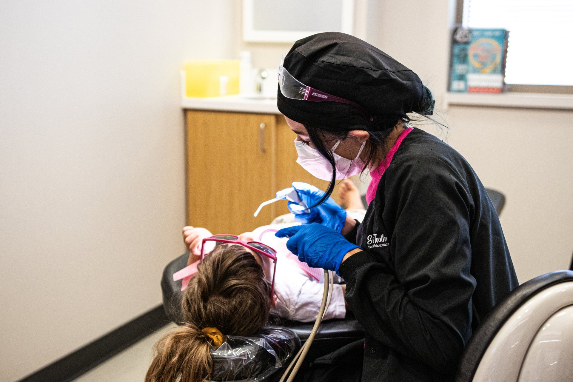 A dental professional in scrubs, mask, and gloves performs a dental procedure on a patient in a clinical exam chair.