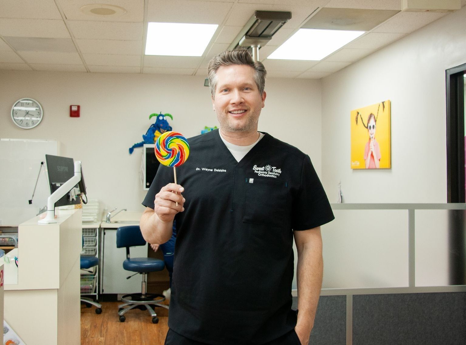 A smiling professional in black scrubs holding a colorful lollipop in a dental or medical office.