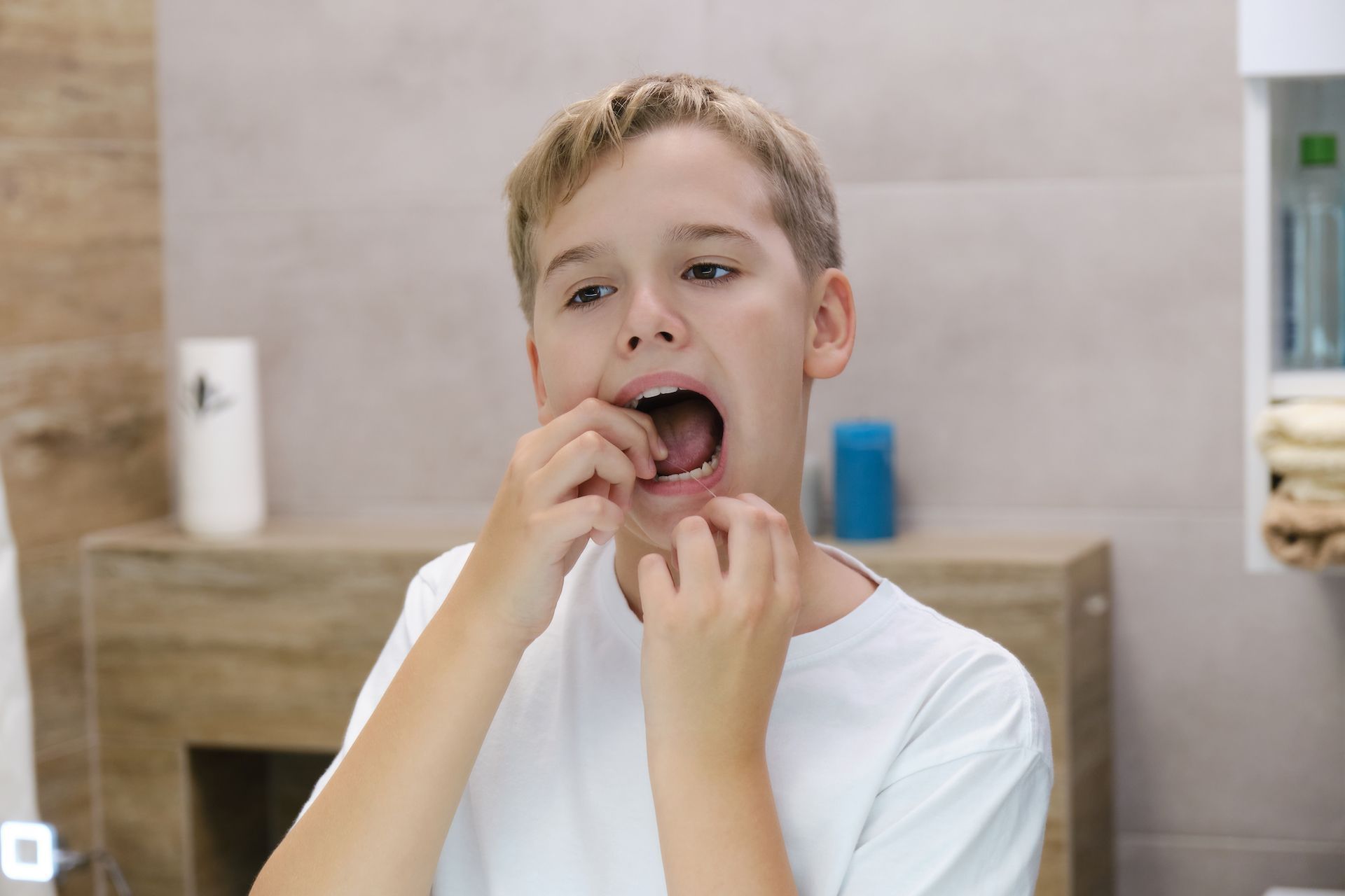 A person in a white t-shirt standing in a bathroom, using their fingers to inspect their teeth while looking in a mirror.