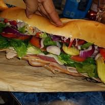 Close-up of a large submarine sandwich filled with various meats, vegetables, and condiments on a light-colored paper.