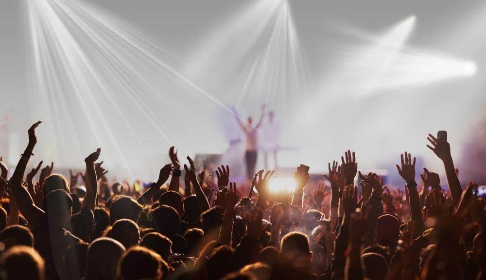Concert crowd with hands raised towards a stage lit by bright spotlights.