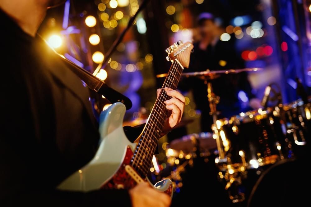 Guitarist playing on stage with a drum set and other musicians in the background; brightly lit.