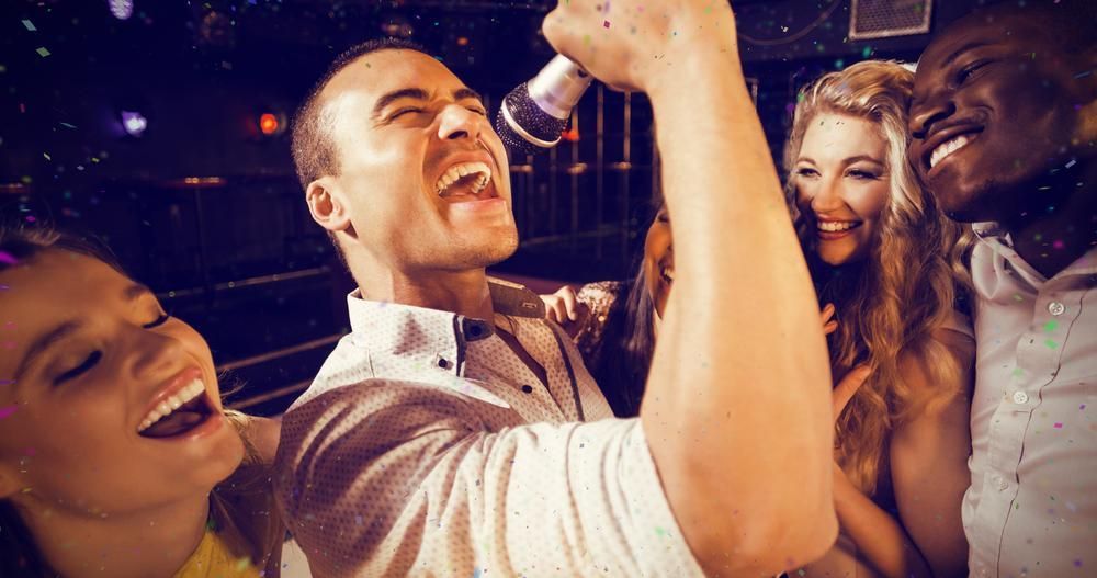 Man singing into microphone, surrounded by laughing friends in a dimly lit bar.