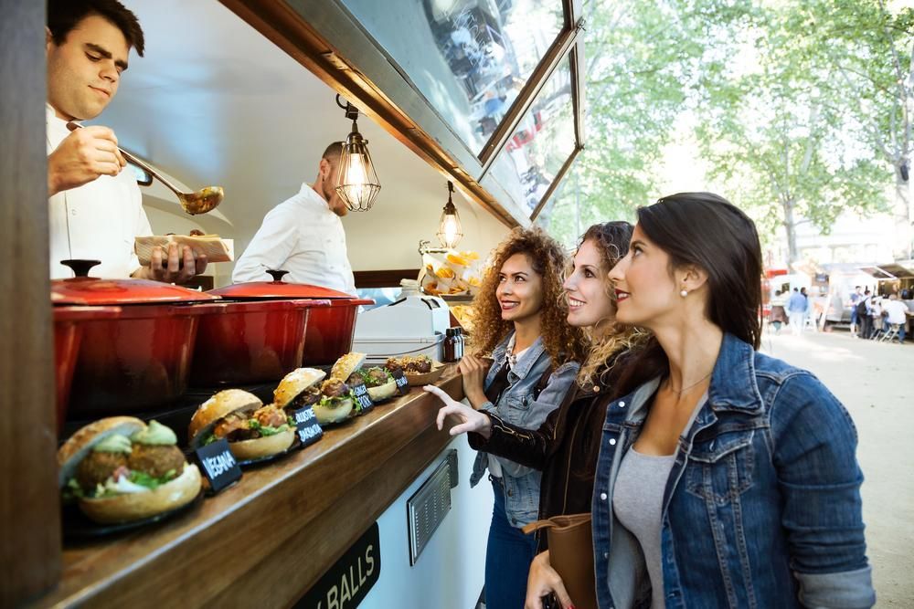 Three women ordering food from a food truck. Workers prepare food, customers point at menu.