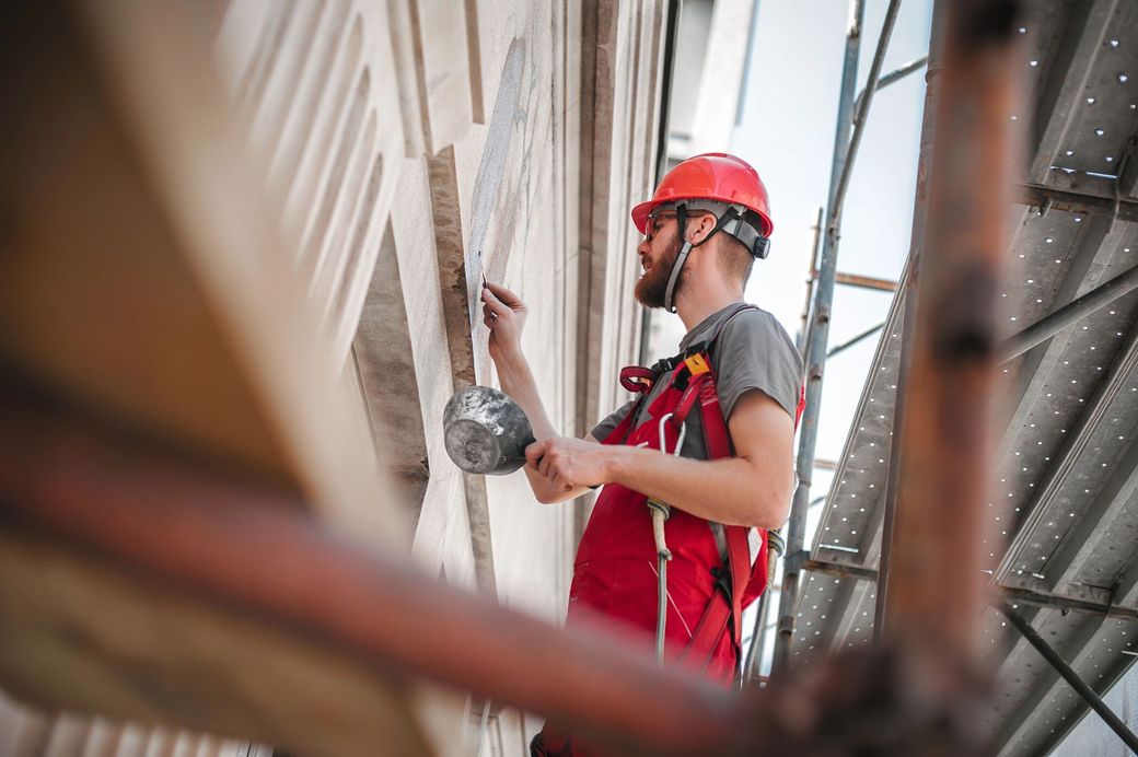 Un operaio edile con elmetto rosso e tuta da lavoro ripara la facciata di un edificio mentre è imbracato a un'impalcatura.