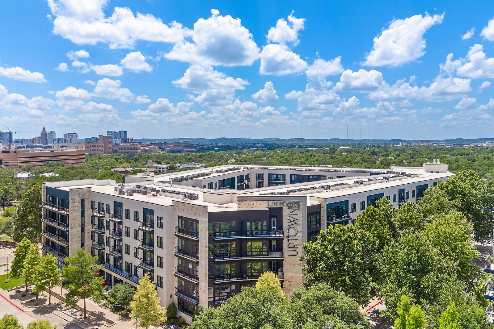 Aerial view of a modern apartment complex surrounded by trees under a bright blue sky with clouds