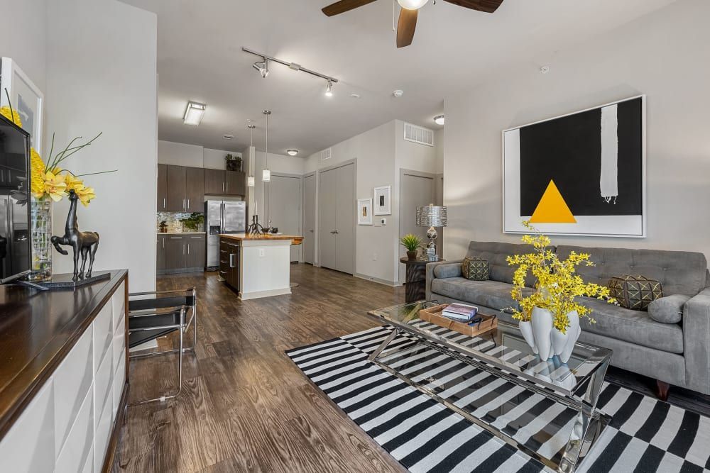 Modern open-concept living room with wooden flooring, featuring a gray upholstered sofa with matching pillows and a large abstract wall art with black, white, and yellow geometric shapes. A glass coffee table with a vase of yellow flowers sits atop a black and white striped area rug, complementing a smaller vase of yellow flowers on a console table by the kitchen. The kitchen area includes wood cabinetry, stainless steel appliances, and a kitchen island with a white countertop and bar stools. The space is illuminated with a combination of track lighting and a ceiling fan at Marq Uptown in Austin, TX.