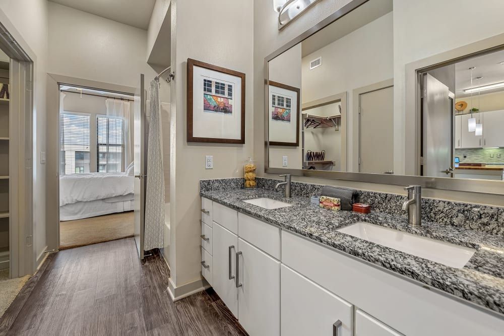 Modern bathroom interior with double sink vanity featuring a granite countertop, several drawers, and two large mirrors reflecting a well-lit bedroom and open shelving. There is a framed picture on the wall, and amenities like soap and sponges arranged on the countertop. The floor is finished with wood-like tiles, and the room is brightly lit with natural and artificial light at Marq Uptown in Austin, TX.