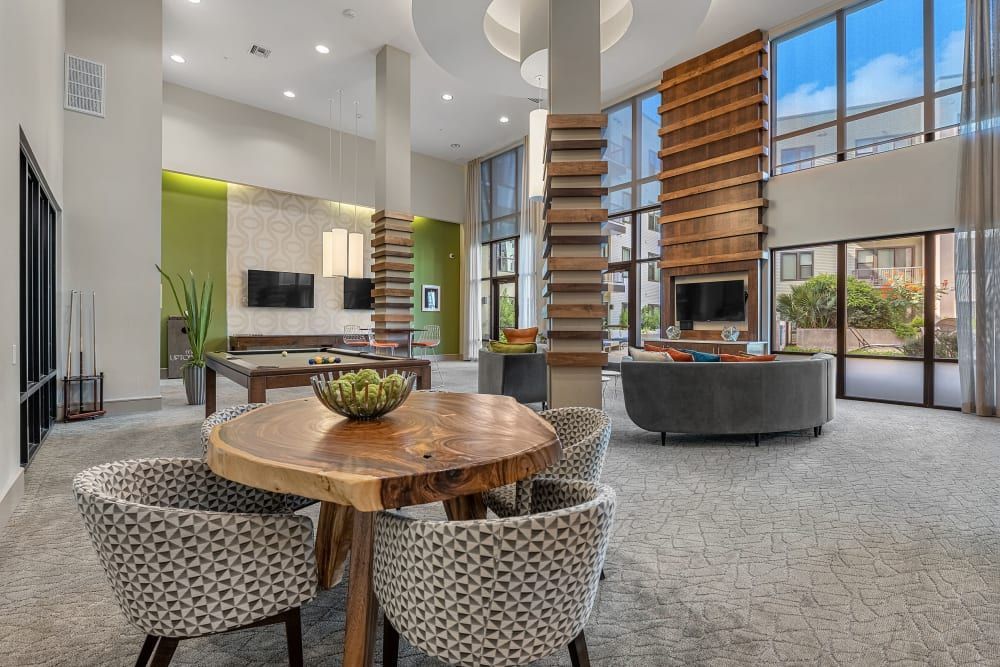 Modern lobby area with high ceilings, large windows, and mixed textures. Features a live-edge wood table with patterned chairs in the foreground, an open staircase with wooden steps to the right, and a cozy sitting area with a flat-screen TV in the background. Decor includes green accent walls, geometric patterns, and indoor plants, complemented by natural light at Marq Uptown in Austin, TX.