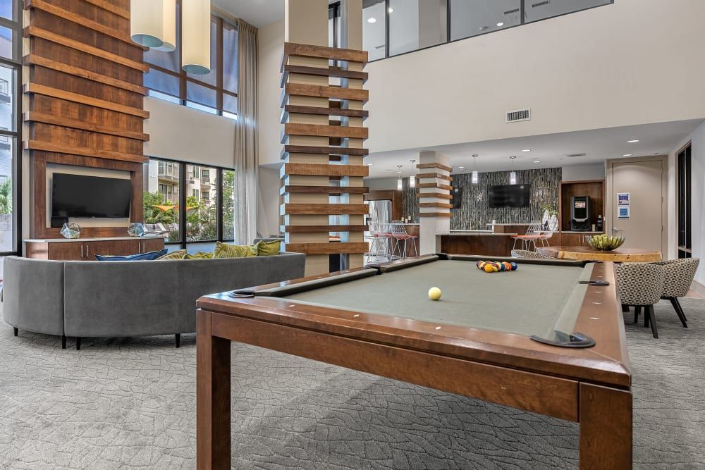 Interior of a modern communal lounge area with high ceilings, featuring a pool table in the foreground with balls ready for a game. Behind it, there is a comfortable seating area with a grey sectional sofa and decorative pillows in front of a flat-screen television. To the right, a kitchenette with bar stools can be seen, and large windows allow for natural light to brighten the room. The space is finished with a combination of wooden accents and a neutral color scheme at Marq Uptown in Austin, TX.