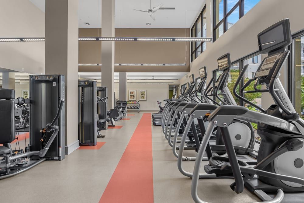 Modern gym interior with a row of elliptical machines on the right, and weight training equipment on the left under bright ceiling lights at Marq Uptown in Austin, TX.