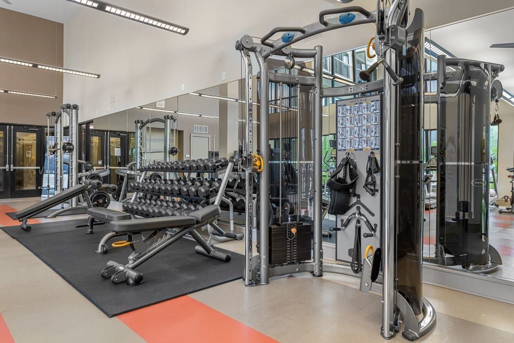 Modern gym interior with a variety of equipment including a dumbbell rack, adjustable weight benches, and cable machines on a red and black rubber floor, reflecting in the mirrors on the wall at Marq Uptown in Austin, TX.