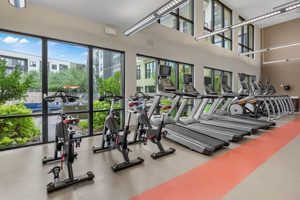 Modern gym interior with a row of treadmills and stationary bicycles facing large windows overlooking green foliage outside, set on a grey floor with a red running track. The high ceiling and ample lighting create a bright and airy exercise space at Marq Uptown in Austin, TX.