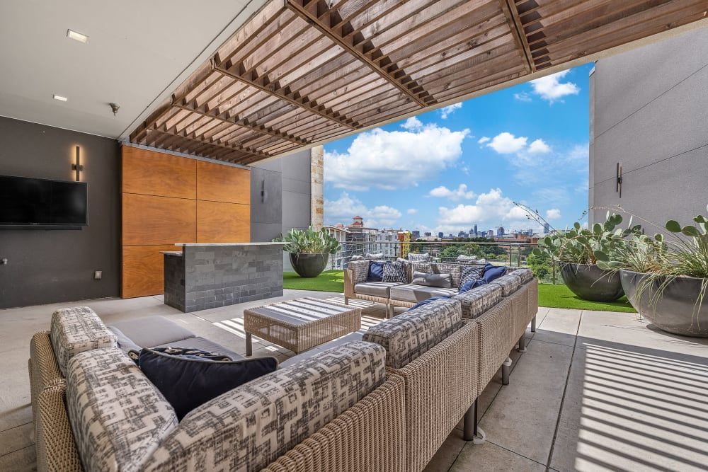 Modern outdoor rooftop terrace with stylish wicker furniture, decorative cushions, and an outdoor kitchen area against a backdrop of a wooden slatted overhang and urban skyline at Marq Uptown in Austin, TX.