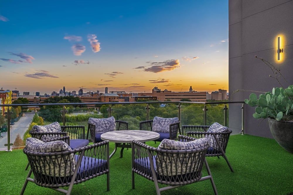 A rooftop terrace at twilight with modern outdoor furniture arranged on artificial grass. The seating includes patterned chairs with cushions around a circular table. A single light fixture illuminates the wall beside a potted cactus plant. In the background, a city skyline is visible under a colorful sunset sky with scattered clouds at Marq Uptown in Austin, TX.