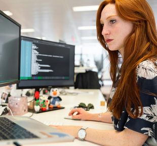 Woman working at a desk with dual monitors in a bright office, typing on a laptop