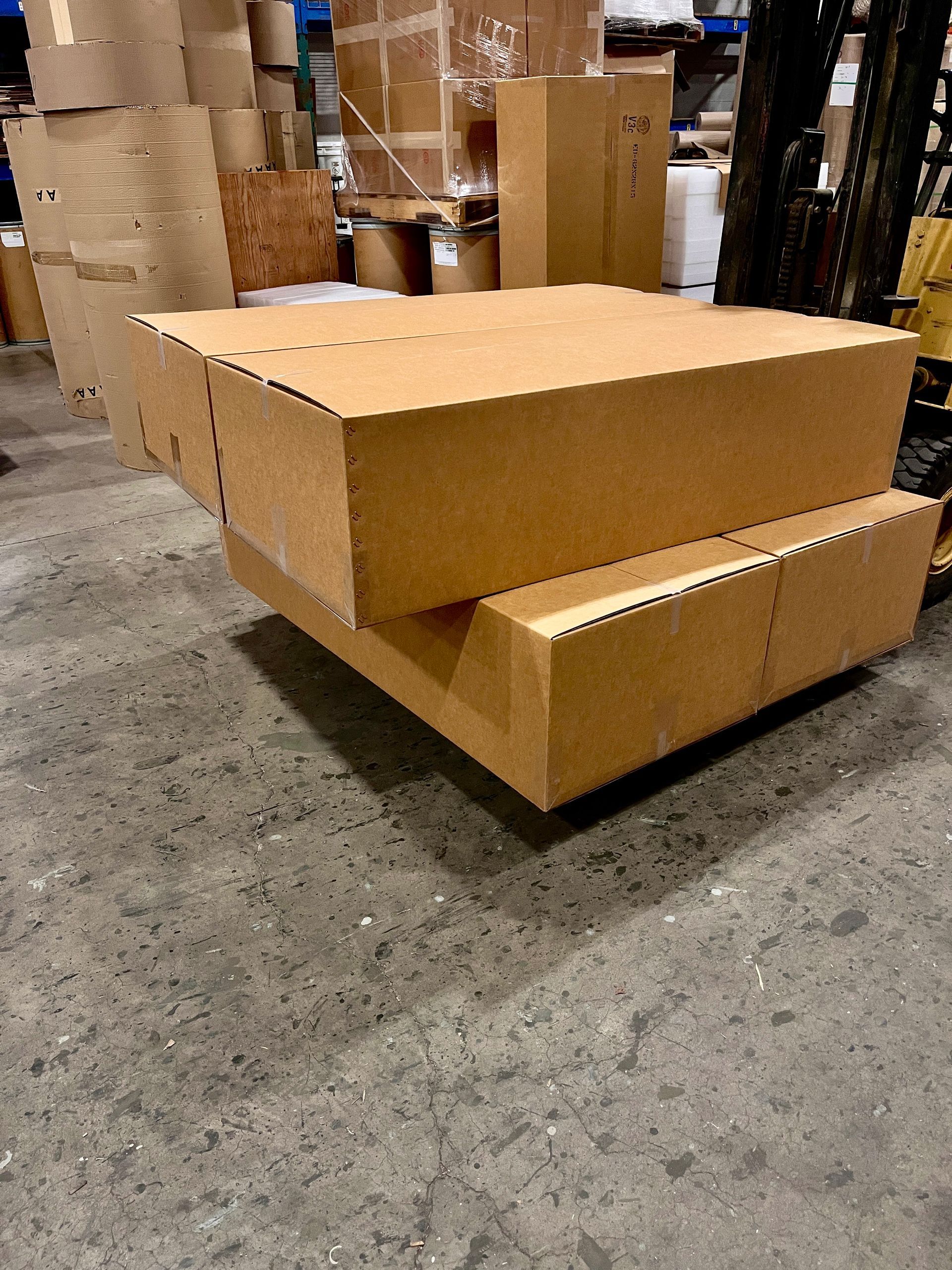 Two stacked cardboard boxes on a warehouse floor. Other boxes are in the background near a forklift.