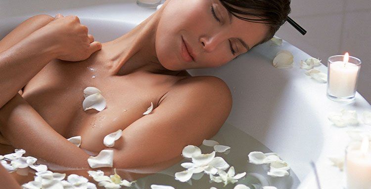Eine Frau liegt in einer Badewanne, umgeben von Blütenblättern und Kerzen.