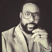 Man with glasses and beard, wearing a blazer and turtleneck, resting chin on hand.
