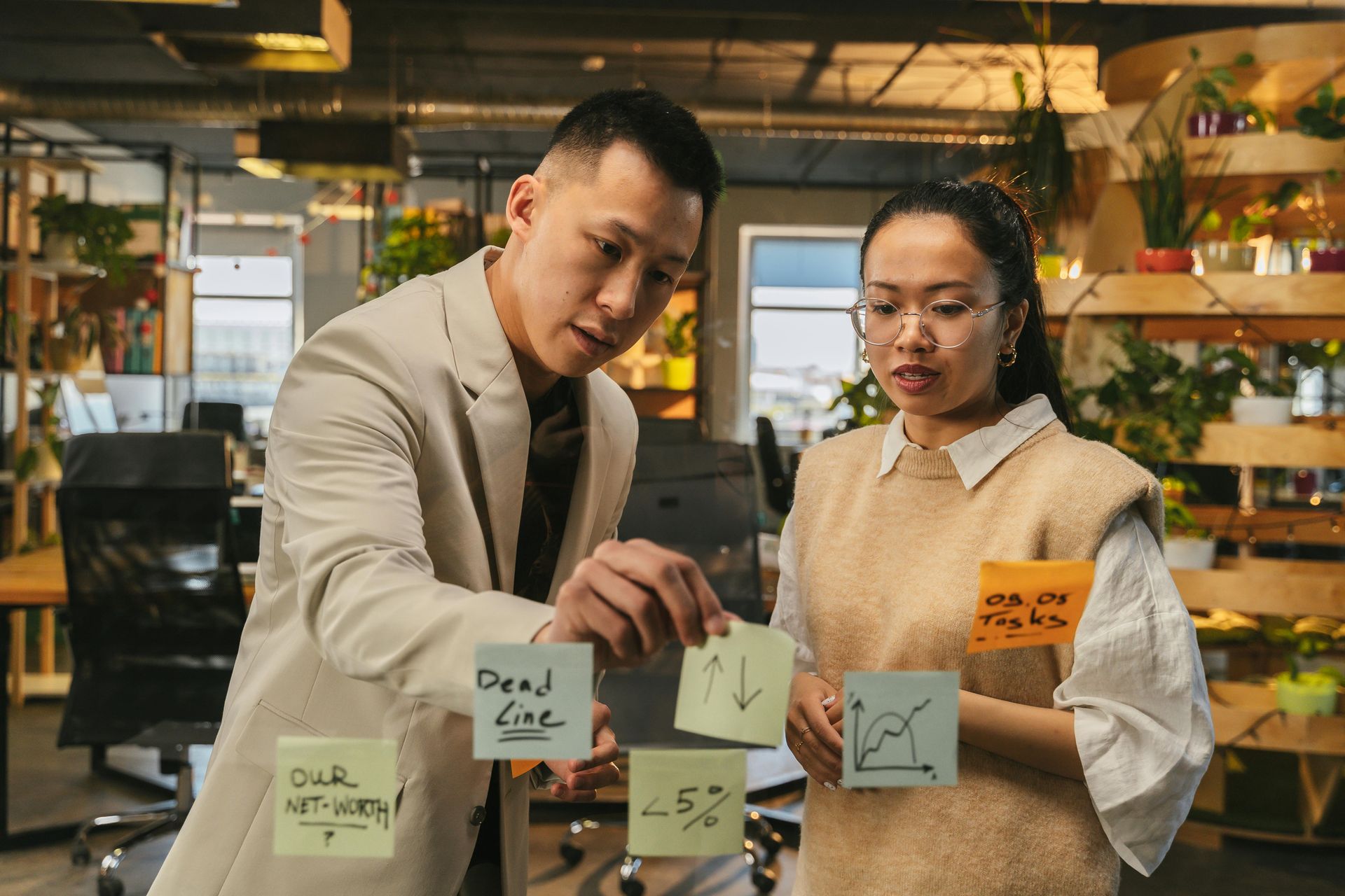 Two people in an office looking at sticky notes on a glass wall, reviewing work tasks.
